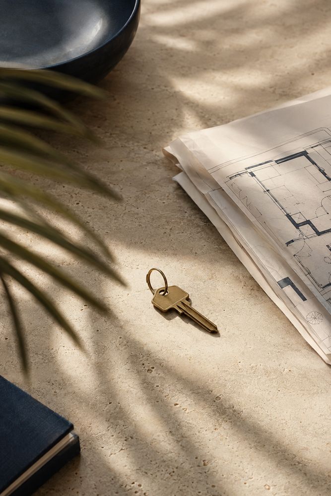 A brass house key on a stone surface beside architectural plans